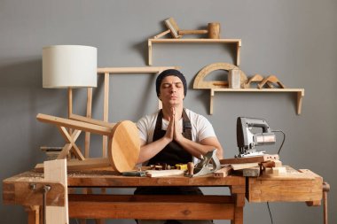 Indoor shot of Caucasian young adult craftsman sitting at table in his workshop, keeping hands in praying gesture, keeps eyes closed, working in joinery, trying to relax with yoga.