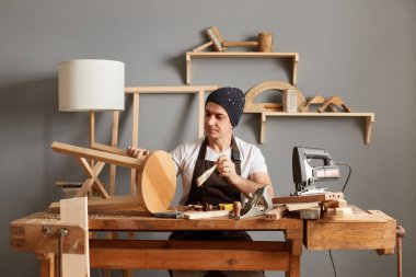 Indoor shot of concentrated man carpenter painting wooden chair made by himself, painting wooden product with varnish, sitting at table in his workshop.