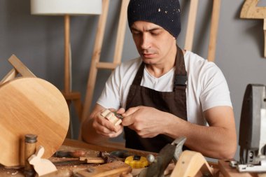 Portrait of carpenter man in brown apron making a handmade wooden toy in a home workshop, handyman carves a toy in wood with a special tool, joiner in the workshop.
