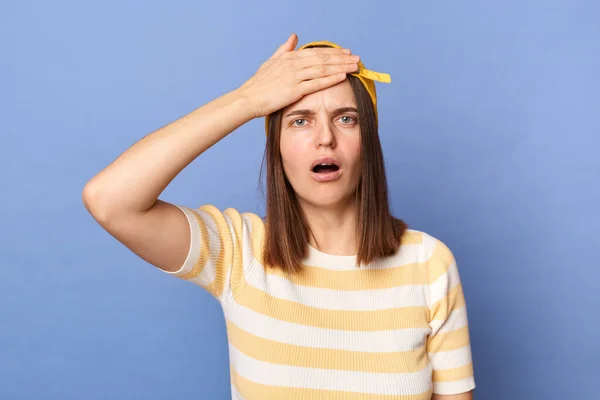 Indoor shot of shocked sad upset teenager girl wearing striped T-shirt and baseball cap, showing facepalm gesture, making mistake, having problems, posing isolated over blue background.