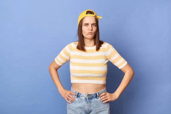 Indoor shot of offended angry teenager girl wearing striped T-shirt and baseball cap posing isolated over blue background, keeps hands on hips, blowing cheeks.