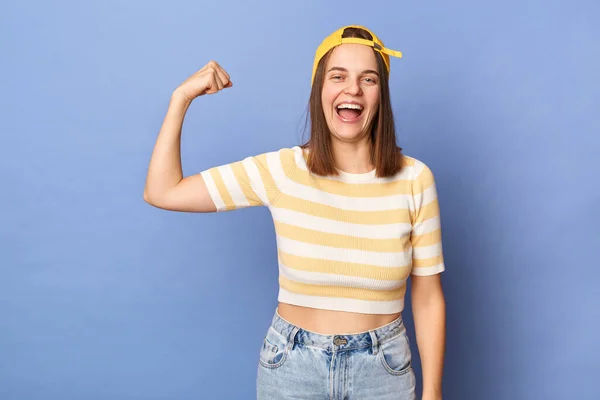 Indoor shot of excited happy smiling teenager girl wearing striped T-shirt and baseball cap posing isolated over blue background, raised her arm. showing her power and biceps.