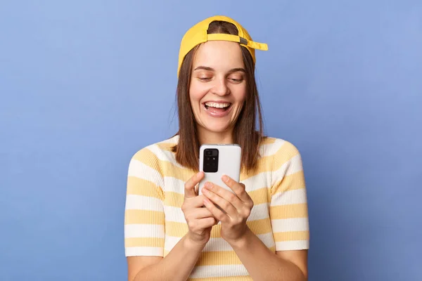 Indoor shot of smiling happy cheerful Caucasian teen girl wearing baseball cap and casual T-shirt standing isolated over blue background, holding smartphone, chatting with friends.