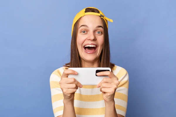 Indoor shot of extremely happy positive optimistic teenager girl wearing baseball cap and stripe T-shirt, holding smart phone, playing video games, winning level, posing isolated over blue background.