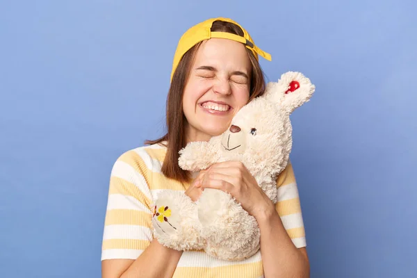 Indoor shot of smiling happy satisfied teenager girl wearing baseball cap and stripe T-shirt posing isolated over blue background, standing hugging soft rabbit toy, keeps eyes closed.
