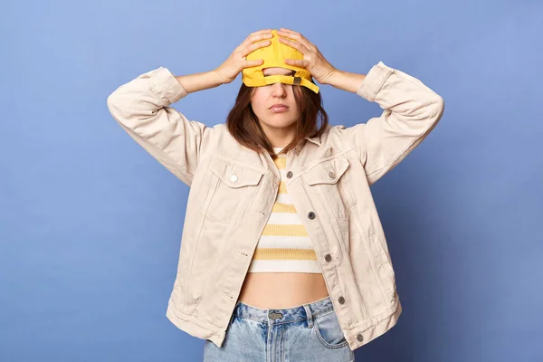 Portrait of sad upset Caucasian teenager girl wearing baseball cap and jacket posing isolated over blue background, pulling her hat on her eyes, expressing sorrow and sadness.