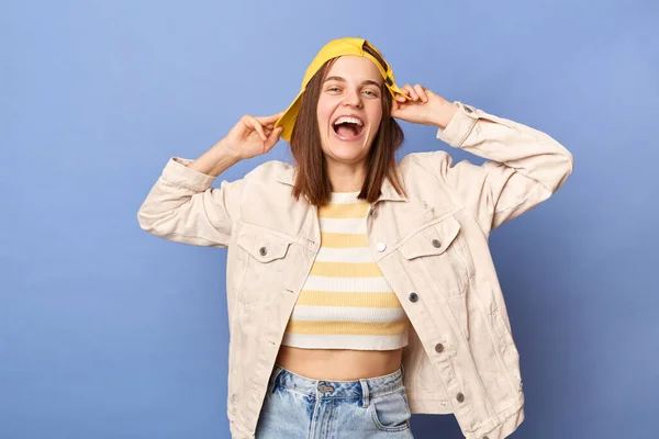 Image of joyful cheerful Caucasian teenager girl wearing baseball cap and jacket posing isolated over blue background, screaming with happiness, keeps hand on hat.