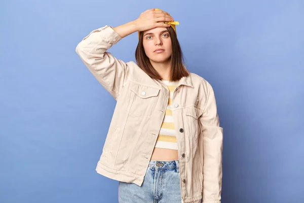 Indoor shot of despair dissatisfied teenager girl wearing baseball cap and jacket posing isolated over blue background, showing facepalm gesture, looking at camera with sad expression, loosing.
