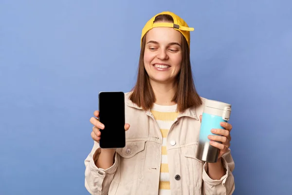 Indoor shot of smiling winsome teenager girl wearing baseball cap and jacket, showing cell phone with empty display, advertisement, drinking coffee, posing isolated over blue background.