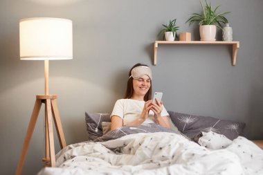 Indoor shot of young woman wearing sleeping mask sitting in bed using mobile cell phone, typing sms message, resting at home, relaxing, expressing positive emotions.