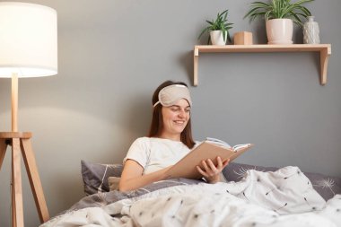 Image of smiling young brown haired woman wearing white t-shirt and blindfold, holding in hand and reading book while rest at home, being involved in interesting plot.