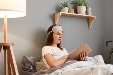 Indoor shot of serious concentrated brown haired woman in white t-shirt and sleeping mask, holding book in hands, reading novel or poems, being at home in bed.