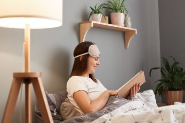 Side view portrait of joyful cheerful young brown haired woman wearing white t-shirt and blindfold, reading interesting book while rest at home, smiling, expressing positive emotions.