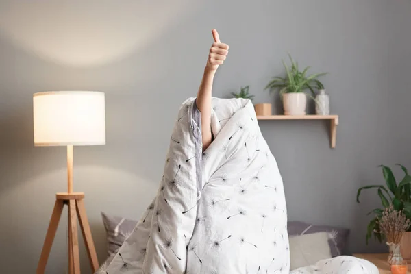 Indoor shot of unknown person sitting on bed wrapped in blanket, hiding her head, showing raised hand and thumb up, demonstrates like gesture, being sleepy, likes wake up early.