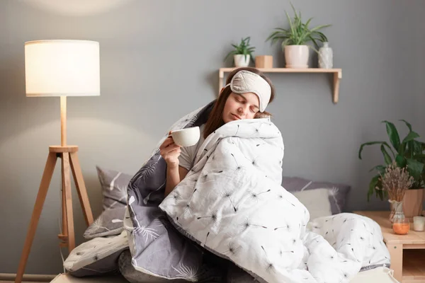Indoor shoot of sleepy woman wearing blindfold sitting on bed wrapped in blanket, holding cup of coffee, having nap, posing with closed eyes, drowsing.