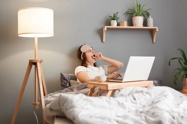 Indoor shot of sleepy bored woman wearing sleeping mask working on laptop from home while lying on bed, spend long hours in front of monitor, feels sleepy, yawning, covering mouth with palm.