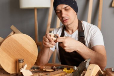 Photo of young adult handyman carpenter wearing white t-shirt, black cap and brown apron, cutting curved piece of wood by wood cutter, making wooden toys.