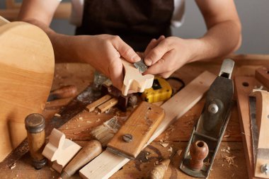 Closeup of man carpenter wearing white t-shirt, black cap and brown apron working, uses chisel for cutting shapes and curves in wood sits at table surrounded by different instruments.