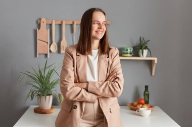 Portrait of smiling attractive young adult woman wearing beige suit standing near table on kitchen at home and looking away, keeps arms crossed, posing at home in the morning.