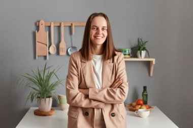 Indoor shot of satisfied delighted brown haired woman wearing beige suit standing near table on kitchen at home, looking at camera, keeps arms folded, wakes up in good mood.