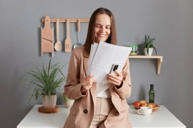 Image of attractive positive brown haired woman wearing beige suit standing near table on kitchen at home, holding smart phone, checking information in documents, enjoying her work.