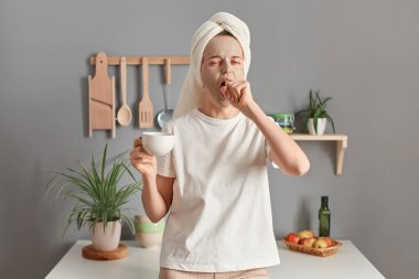 Portrait of young yawning woman in towel wrapped around hair, with morning cup of coffee, standing in kitchen, sleepy tired girl in white T-shirt and cosmetic mask on face drinking coffee.
