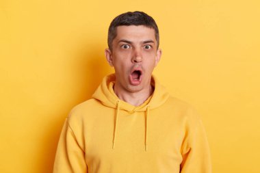 Portrait of surprised man with dark hair wearing casual style hoodie standing isolated over yellow background, sees something astonishing, keeps mouth widely opened.