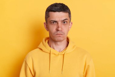 Portrait of annoyed frustrated man with dark hair wearing casual style hoodie standing isolated over yellow background, looking at camera with frowning face, being aggressive.