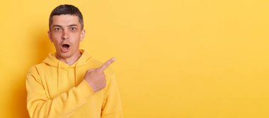 Portrait of amazed surprised man with dark hair wearing casual style hoodie, pointing away at copy space, standing isolated over yellow background, showing advertisement area for promotional text.