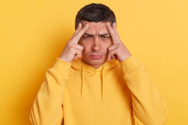 Unhappy frustrated man wearing casual style hoodie, massaging temples feeling headache, suffering migraine or high blood pressure, trying to concentrate, standing isolated over yellow background.