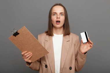 Indoor shot of shocked woman with brown hair wearing beige jacket posing isolated over gray background, holding clipboard and credit card, having problems with bank account.
