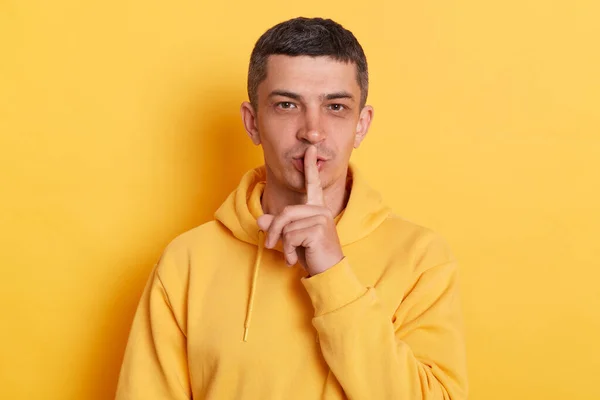 Shh, be quiet. Portrait of man with dark hair wearing casual style hoodie standing isolated over yellow background, showing silence gesture with finger on her mouth, asking to stay quiet, keep secret.