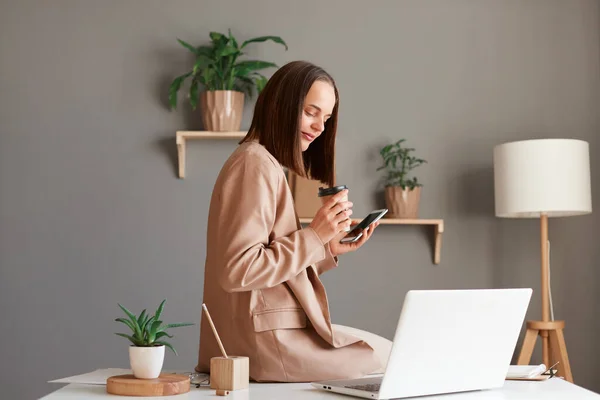 Indoor shot of smiling positive Caucasian dark haired woman in beige suit sitting on table in office with smart phone and takeaway coffee, working on laptop.