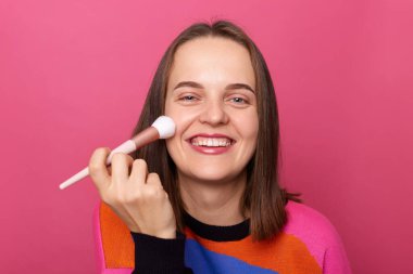 Photo of young happy smiling beautiful woman doing contouring apply blush on cheeks isolated over pink background, wearing colorful sweater, having brown hair, looking at camera, enjoying visage.