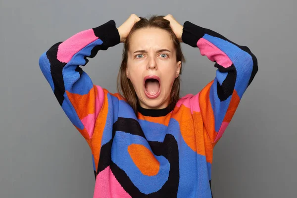 Indoor shot of despair woman with brown hair wearing sweater standing isolated over gray background, screaming loudly, expressing rage, keeps mouth widely opened, pulling her hair.