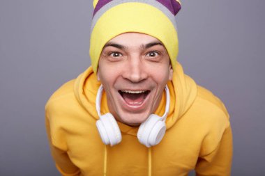 Closeup portrait of excited overjoyed hipster man wearing yellow hoodie and beanie hat standing against grey wall expressing happiness, celebrating sudden success, looking at camera, screaming wow.