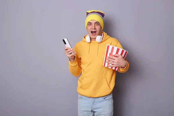 Image of glad cheerful man wearing hat and hoodie having fun, holding popcorn bucket in cinema, enjoys funny movie, standing against gray, using mobile phone, screaming, expressing surprise and shock.