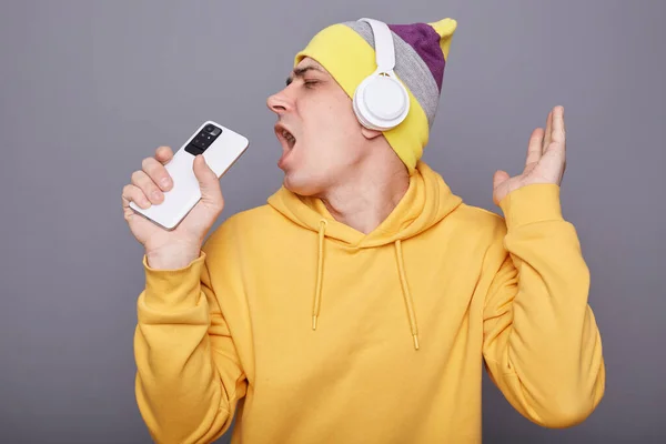 Indoor shot of cheerful satisfied man in yellow hoodie singing song and using smart phone as microphone, having fun, wearing wireless headset, dancing isolated over gray background.