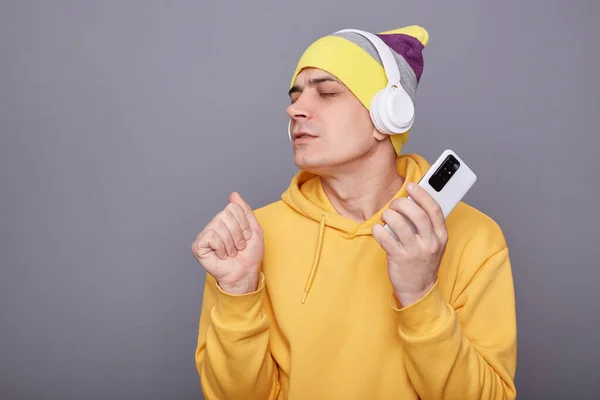 Portrait of overjoyed Caucasian guy singing song and holding smart phone in hands, clenched fist, enjoying song, keeps eyes closed, wearing wireless headset, dancing isolated over grey background.