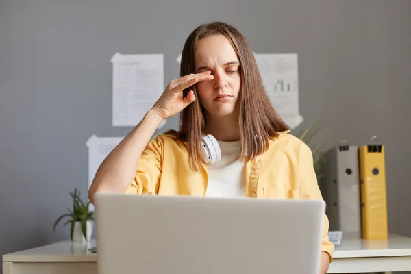 Image of exhausted woman student or freelancer with brown hair, female working on laptop during long hours, feels fatigue, being overworked, rubbing her tired eyes, office background.