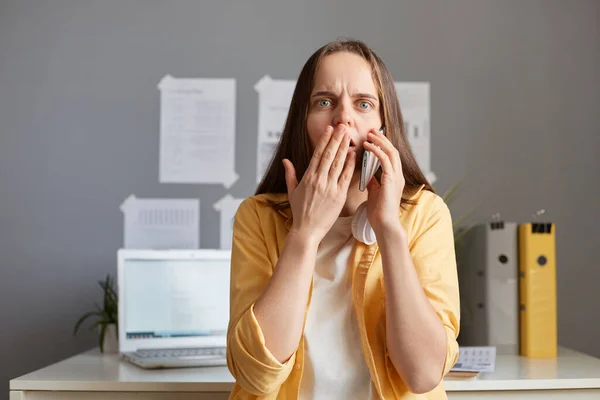 Indoor shot of shocked astonished Caucasian woman with brown hair sitting in office at her workplace and talking on mobile phone, wearing yellow shirt, hearing surprised news, covering mouth with palm.