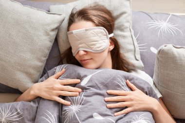 Portrait of calm young woman wearing sleeping mask lying in comfortable bed on pillow and sleeping, female in blindfold resting, lying under blanket.