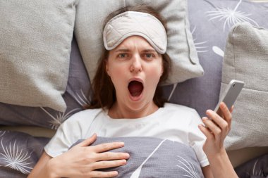 Portrait of shocked surprised young woman wearing sleeping mask lying in bed on pillow, looking at camera with open mouth, screaming, holding cell phone, reading surprised news.