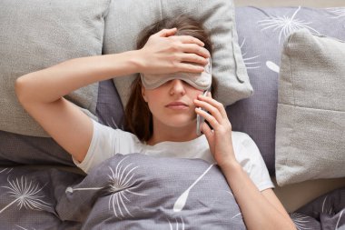 Indoor shot of tired sick unhealthy young woman wearing sleeping mask lying in comfortable bed on pillow and talking on smart phone, keep hand on forehead, feels oversleep.