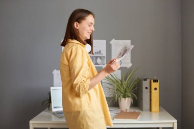 Side view portrait of hard working woman with brown hair wearing yellow shirt posing in office, holding paper documents, standing near table with paper folder and laptop.