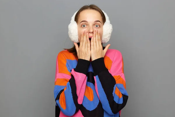 Wow. Photo of young woman wearing white fluffy ear warmers and colorful sweater, feeling shocked and deeply surprised, posing isolated on gray background, covering mouth with palms.