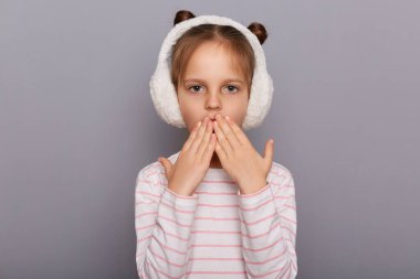 Portrait of shocked scared little girl wearing fur earmuffs and striped shirt, looking at camera with surprised facial expression, covering mouth with palms, standing isolated over gray background.