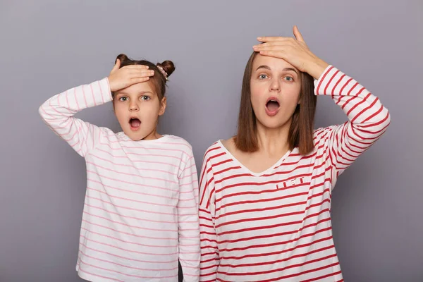 Indoor shot of scared shocked woman and little girl with hair buns wearing casual clothes standing isolated over gray background, looking at camera with frighten expression.