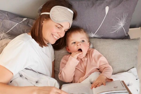一个漂亮的年轻母亲带着她的小女儿躺在床上看书 表达着幸福 在睡前为迷人的婴儿读书的横向镜头 — 图库照片