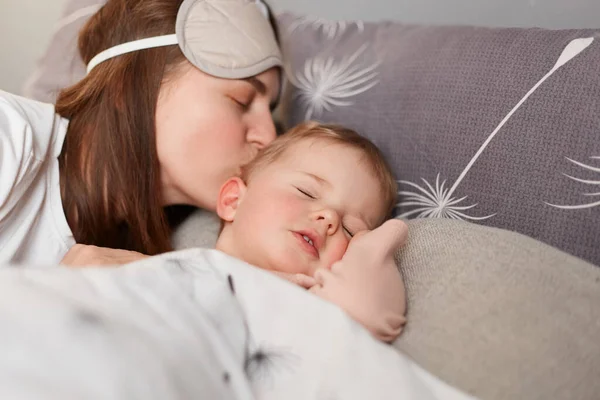 年轻的母亲黑头发的女人吻着甜美的小宝宝 可爱的孩子躺在妈妈身边睡觉 而父母吻着她 家的内部 养育子女 — 图库照片